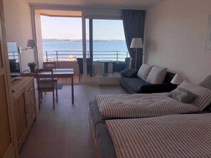 A hotel room with a bed, a sofa, a desk, and a chair. There is a balcony with a view of the sea.