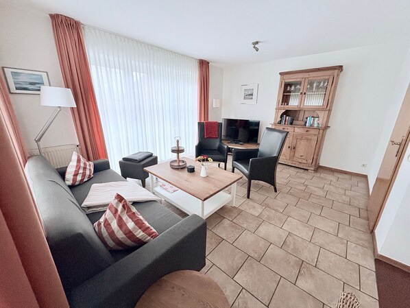 Un salon moderne avec un canapé, des fauteuils, une table basse et un téléviseur. On y trouve un meuble en bois avec des portes en verre et une lampe sur une table d’appoint.