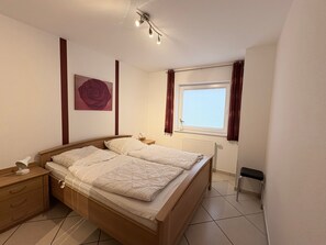 Ein Schlafzimmer mit einem Holzbett, einem Nachttisch, einer Nachttischlampe, einem Heizkörper, einem Fenster mit Vorhängen und einer Wandleuchte.