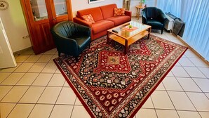 Un salón con un sofá rojo, un sillón negro, una mesa de centro, un tapete estampado, un cuadro en la pared y una ventana con cortinas.