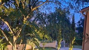 A well-maintained garden with various trees and shrubs, a paved pathway, and a building with a lighted entrance. - Homerez - Villa in Ribérac (Ribérac)