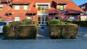 A red-brick building with a central glass door entrance, surrounded by neatly trimmed hedges and a paved driveway. - Achtern 8 - 70018, Germany (Wangerland)