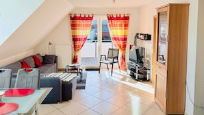 A modern living room with a sofa, ottoman, and a dining table with red placemats. - Achtern 8 - 70018, Germany (Wangerland)