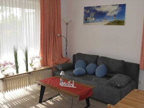 Un salon avec un canapé gris, une table basse en bois recouverte d’un tissu rouge et un tableau représentant un phare accroché au mur.