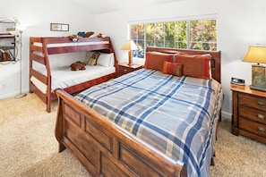 A bedroom with a bunk bed, a large bed with a plaid bedspread, a bedside table with a lamp, and a window with a view of trees. - Beautiful Townhouse! Walking Distance to Bear Mtn and Golf Course! Walk to Hiking! (Big Bear Lake)