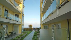 A narrow pathway between two yellow buildings with balconies, a small seating area with chairs, and a clear sky. - Dune Residence Bansin - Apartment B 03 (Heringsdorf)