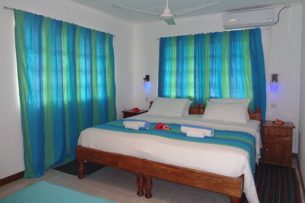 A bedroom with a bed, curtains, a ceiling fan, an air conditioning unit, and a bedside table with a lamp.