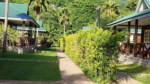 A pathway leading to a resort with green-roofed buildings, palm trees, and lush greenery. - Chez Michelin Pension Residence (La Digue)