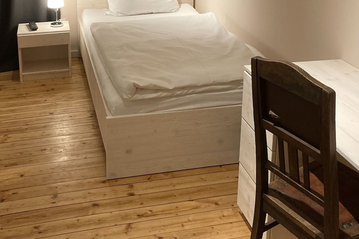 Quarto com cama, mesa de cabeceira com abajur, cadeira de madeira e escrivaninha.
