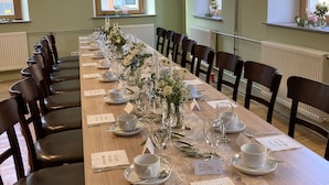 A long dining table set for a meal with chairs on both sides, in a room with green walls and windows. - BürgerhausLöwen (Philippsburg)