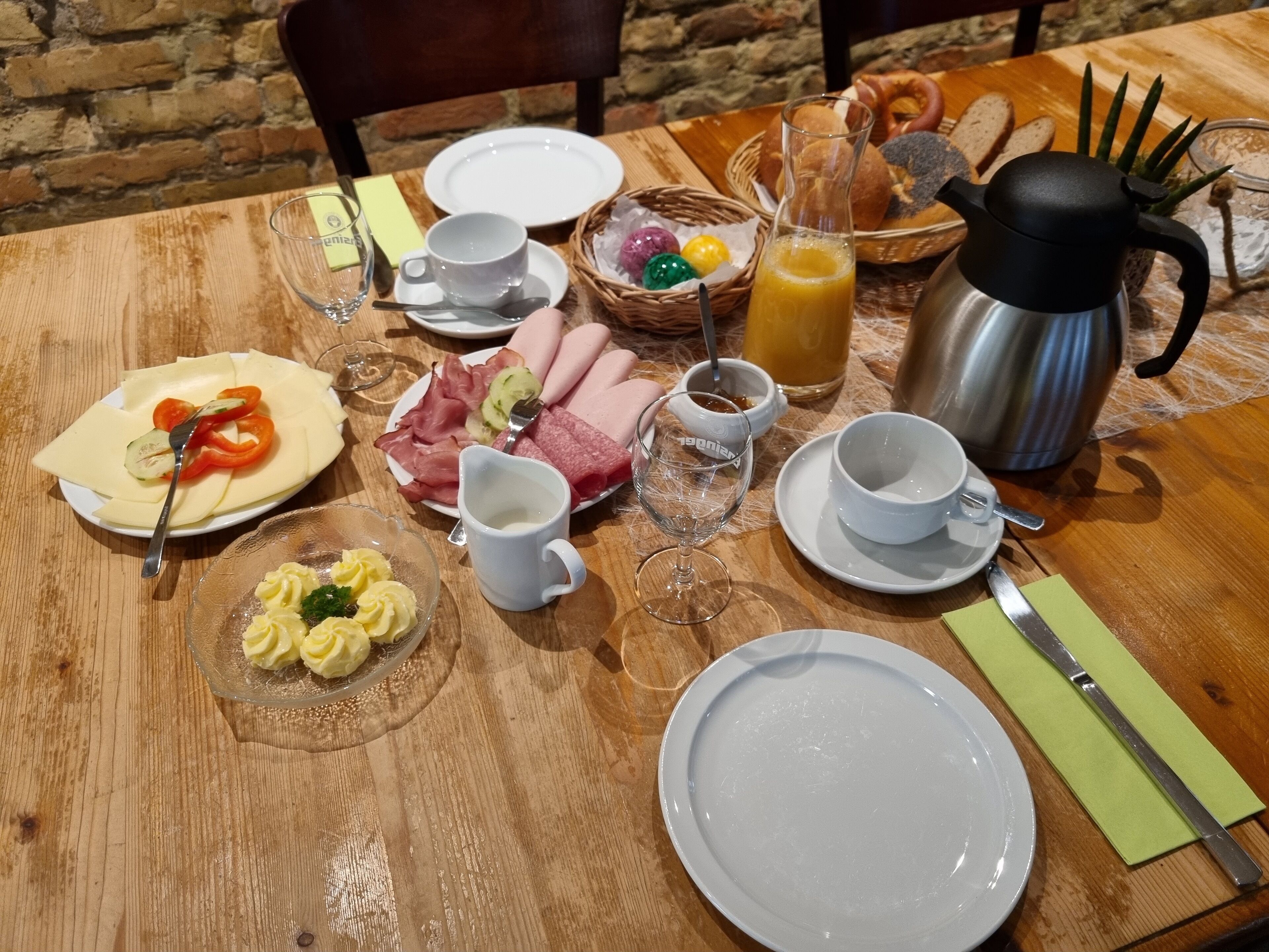 Una mesa de madera con platos, cubiertos, una cafetera y diversos alimentos para el desayuno.