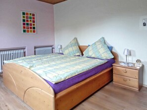 A single bed with a wooden frame, a purple bedspread, and two matching pillows. There is a nightstand with a lamp and a small clock. A radiator is visible on the wall, and a colorful artwork is hung above the bed. - Vacation apartment Beate Engelhardt (Edertal)