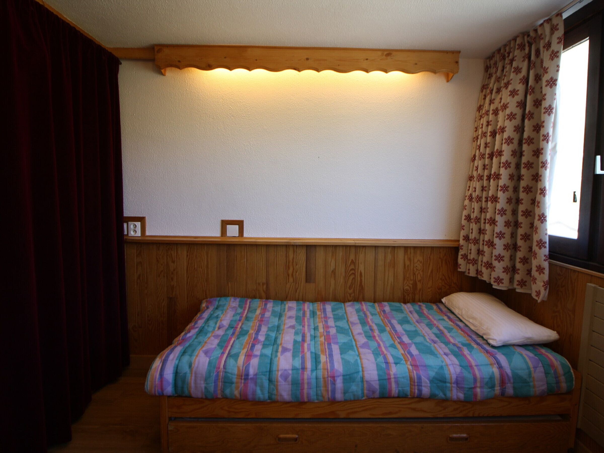 Une petite pièce chaleureuse, avec un lit en bois, une simple tête de lit, une petite étagère et une fenêtre avec des rideaux.