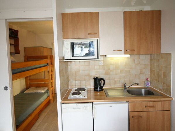A compact kitchen with a microwave, stove, sink, and a small refrigerator. There is a bunk bed in the adjacent room.