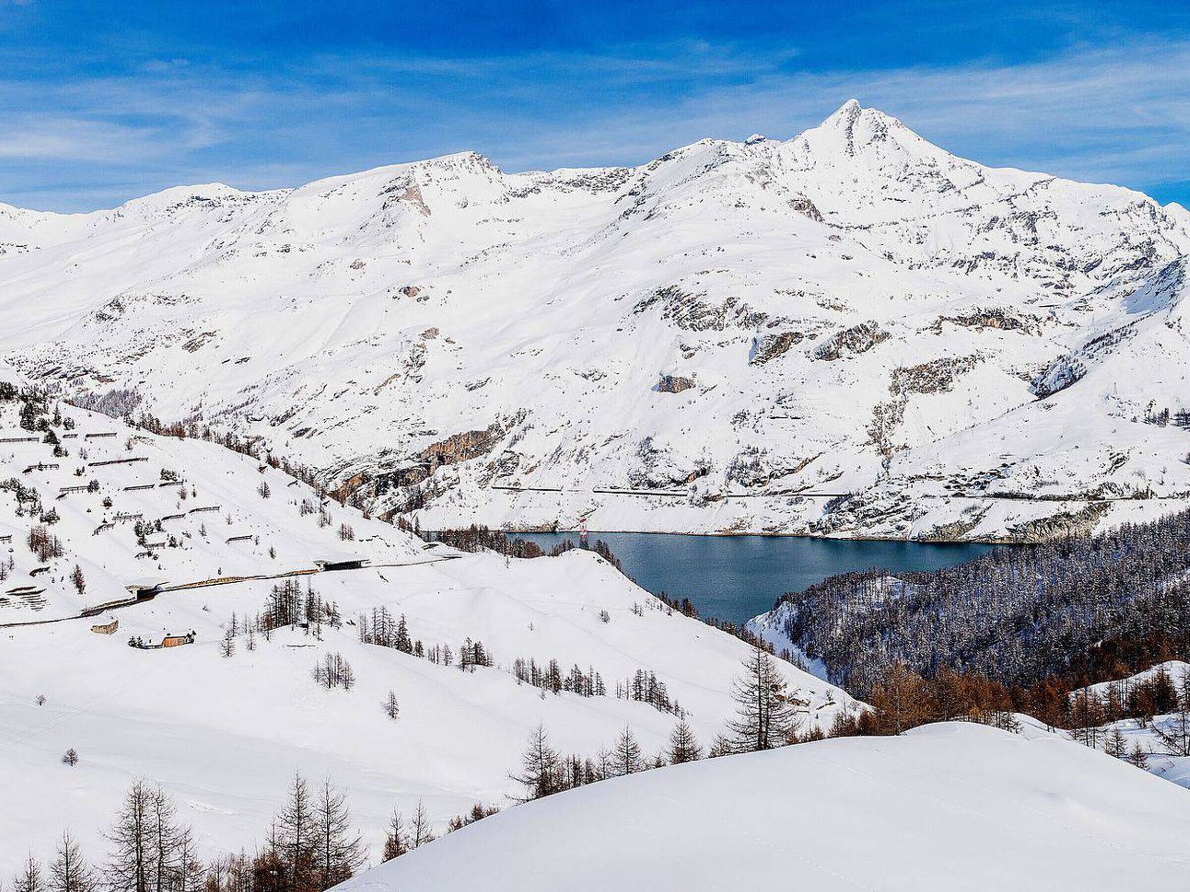 A snow-covered mountain landscape with a lake and a few buildings in the foreground.