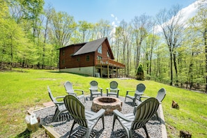 A cabin with a deck, surrounded by a fire pit and Adirondack chairs, set in a forested area.