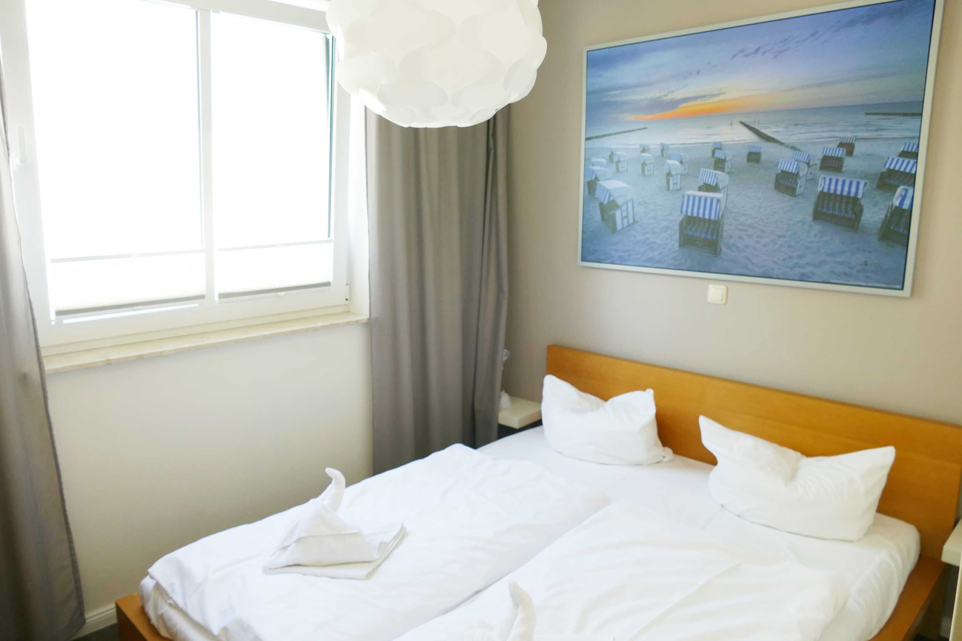 Quarto de hotel com uma cama grande, um quadro de uma paisagem praiana e uma janela com cortinas.