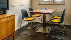 En matplats med ett bord och fyra stolar, en tv placerad på ett träskåp och ett fönster med persienner.