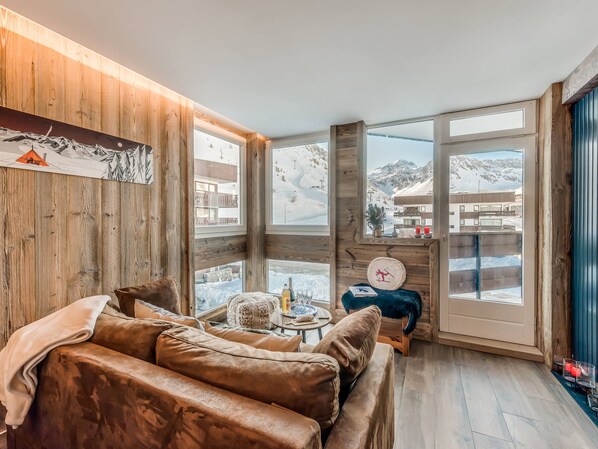 Um quarto acolhedor com parede de madeira, um sofá de couro marrom, uma mesinha com uma garrafa e vista para montanhas nevadas através de grandes janelas.