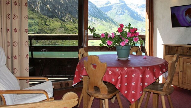 A room with a table set for two, a wooden chair, a sofa, and a view of mountains through large windows. - Large Studio for 4 - Lake View in Tignes (Tignes)