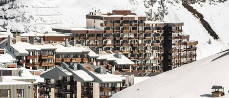 A snow-covered mountain town with modern multi-story buildings, some with balconies, and a ski slope in the background.