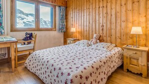 A cozy room with wooden paneling, a bed with floral bedding, a desk with a chair, a teddy bear, and a window with a view of mountains. - Apartment 3*, Savoyard style, 4 ppl, renovated, lake (Tignes)