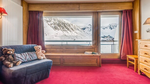 A room with a large window offering a view of snow-covered mountains, a blue sofa with two teddy bears, a wooden bench, and a red carpet. - Apartment 3*, Savoyard style, 4 ppl, renovated, lake (Tignes)