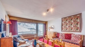 A cozy room with a wooden dining table set for two, a sofa with patterned cushions, and a large window offering a view of a snowy mountain. - Apartment 3*, Savoyard style, 4 ppl, renovated, lake (Tignes)