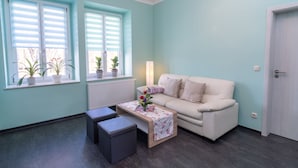 Ein Zimmer mit blauer Wand, einem weißen Sofa, einem kleinen Tisch mit floralem Tischläufer, zwei grauen Poufs und einer Tür.
