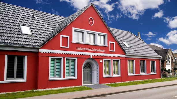 Ein rotes Gebäude mit einem Satteldach, einem zentralen Eingang mit einer Tür und einem Schild darüber.