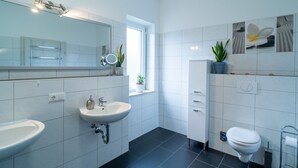 A modern bathroom with a large mirror, two sinks, a toilet, and a window with plants.
