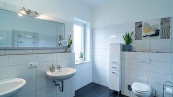 Ein modernes Badezimmer mit einem großen Spiegel, zwei Waschbecken, einer Toilette und einem Fenster mit Pflanzen.