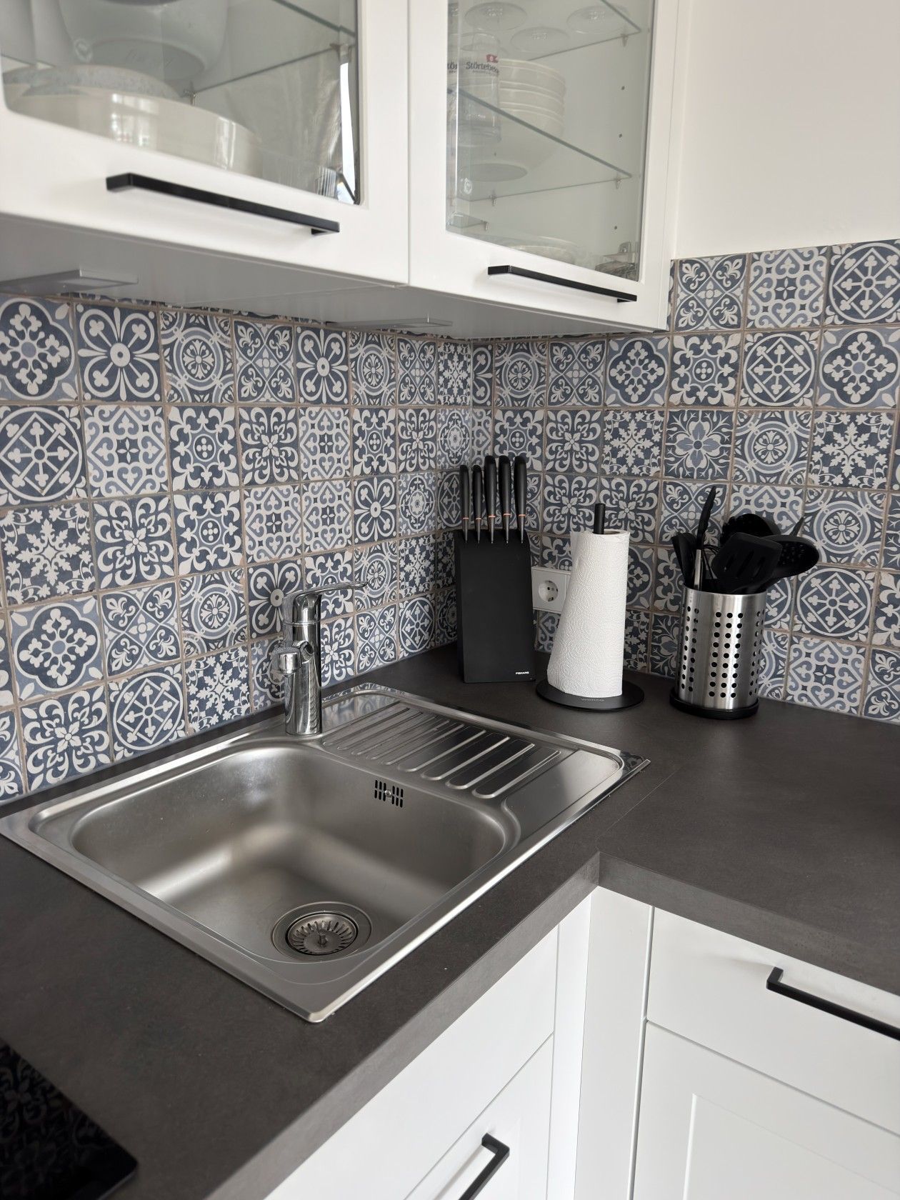 A kitchen with a stainless steel sink, white cabinets, and patterned tile backsplash.