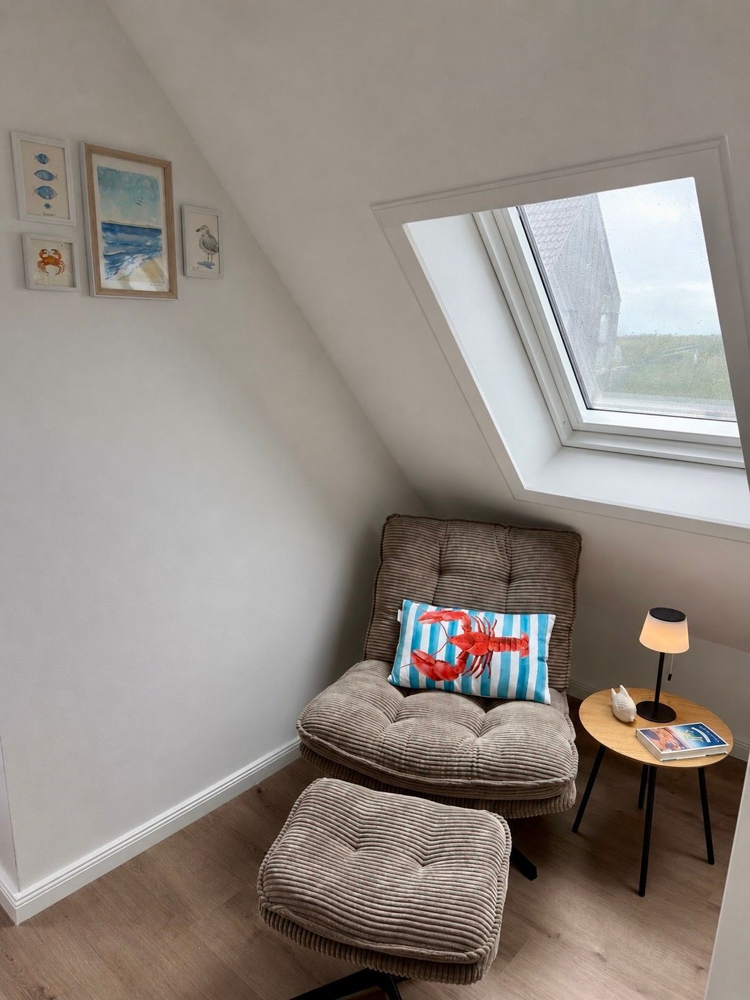 A cozy room with a sloped ceiling, a cushioned chair with a striped pillow, a small round table with a lamp, and a window with a view of the sky.