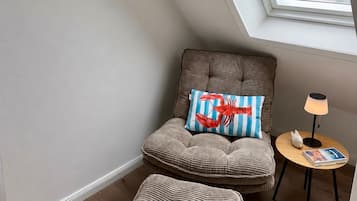 A cozy room with a sloped ceiling, a cushioned chair with a striped pillow, a small round table with a lamp, and a window with a view of the sky.