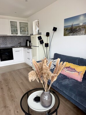 Een moderne keuken met witte kasten en een witte koelkast, een zithoek met een blauwe bank en een vaas met gedroogde planten, en een schilderij van een strandgezicht aan de muur.