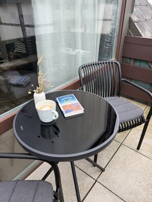 Een ronde tafel met een boek en een kopje, een stoel met een kussen, en een raam met uitzicht op een gebouw.