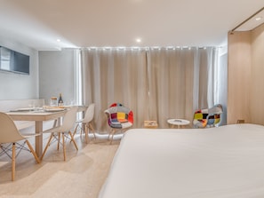 Une chambre d’hôtel moderne dotée d’un grand lit, d’une table à manger avec des chaises, d’un téléviseur fixé au mur et offrant une vue sur la ville par la fenêtre.