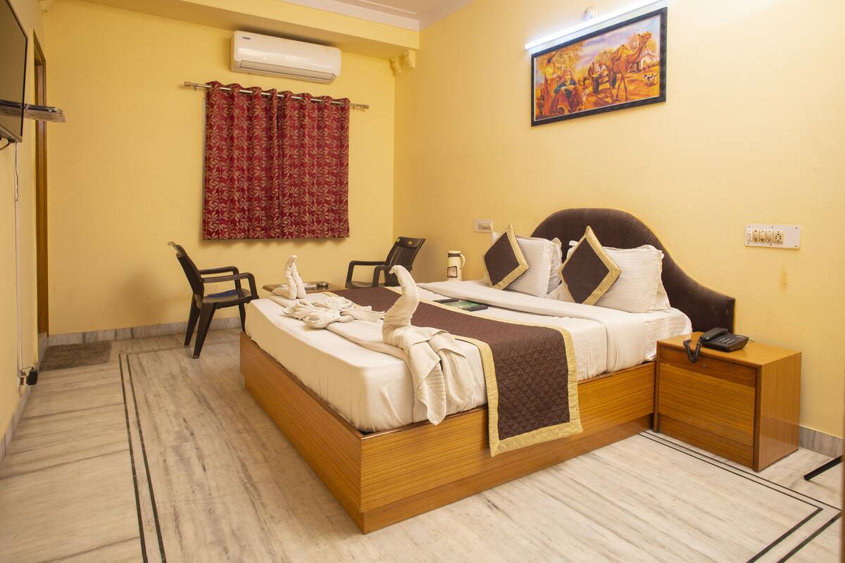 A hotel room with a bed, two chairs, a bedside table with a phone, an air conditioner, a curtain, and a painting on the wall.