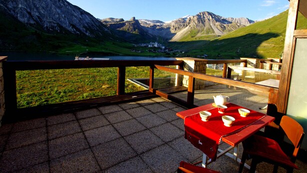 Une terrasse avec une table et des chaises, d’où l’on peut admirer un paysage montagneux avec un lac et des bâtiments visibles au loin.