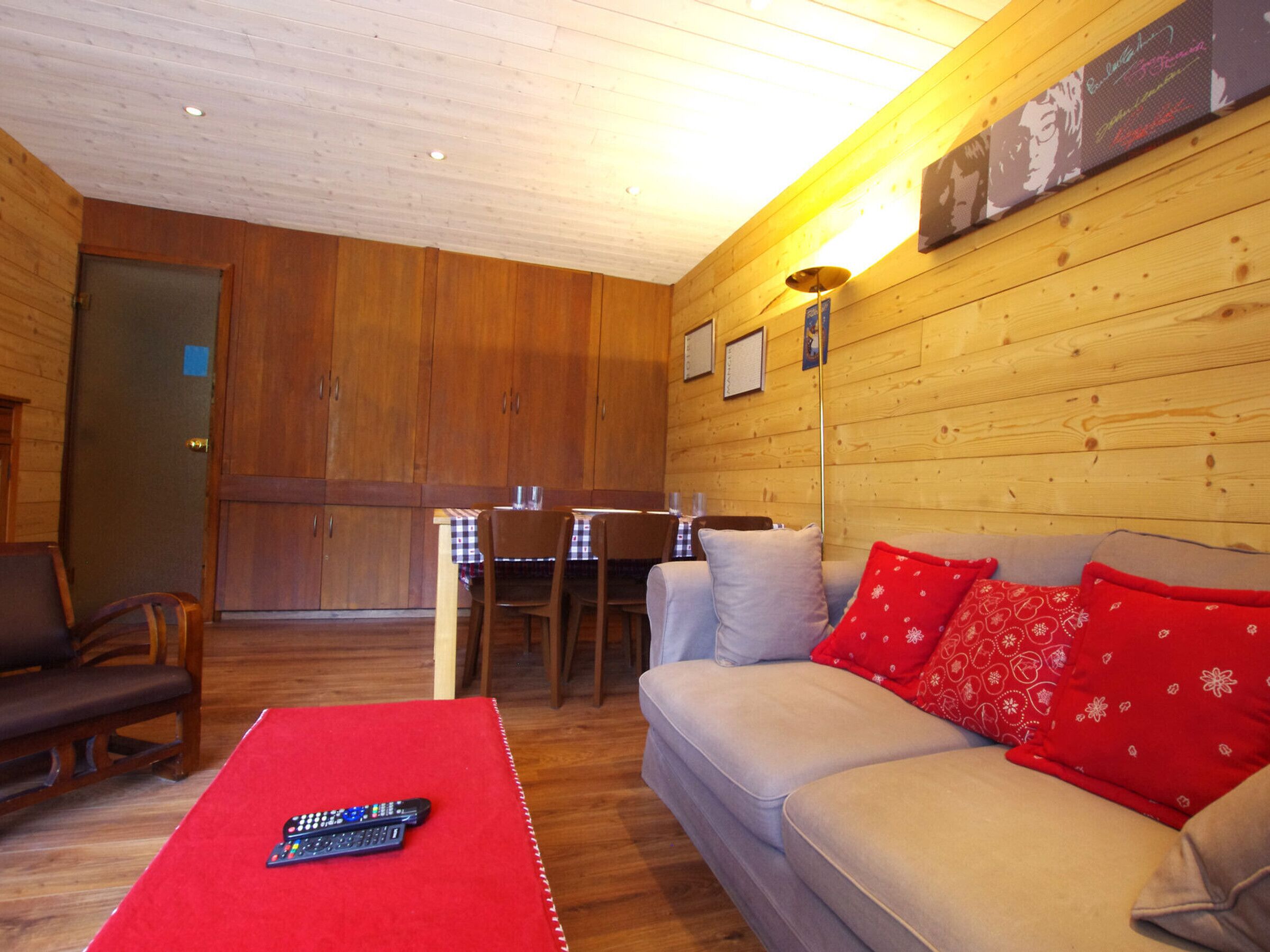 A cozy room with a wooden interior, a sofa with red cushions, a table with a red tablecloth, and a remote control on the table.