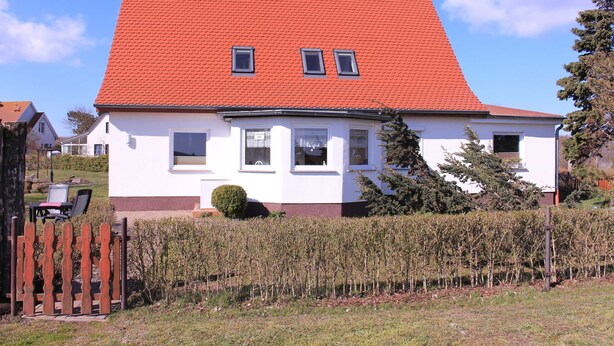 A white house with a red roof, two small windows, and a wooden fence in the foreground.