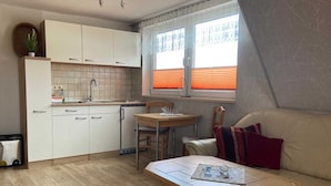 A compact kitchen with white cabinets, a wooden dining table, and a window with orange blinds.