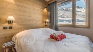 Ein Hotelzimmer mit einer Holzwand, einem großen Bett, zwei Wandleuchten, einem Fenster mit Bergblick und einem kleinen Holzschemel mit einer Zeitschrift.