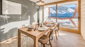 Ein Essbereich mit einem Holztisch, gedeckt für vier Personen, einer hängenden Leuchte und einem großen Fenster mit Blick auf schneebedeckte Berge.