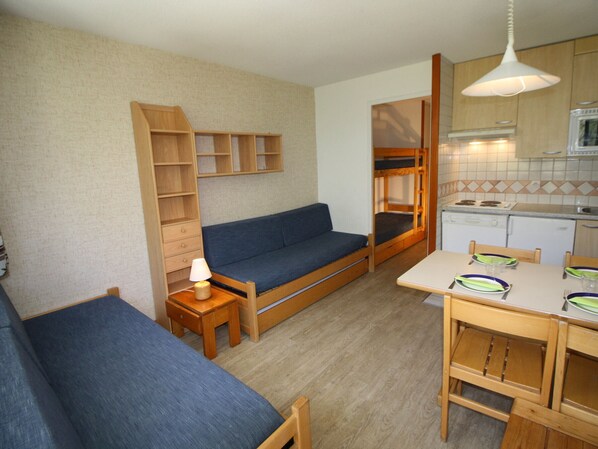 A compact living space with a sofa, a small table, a kitchenette, and a wooden shelf unit.