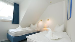 A hotel room with two beds, a window with blue curtains, and a city view. - App. Feusser - at Wenningstedter Strand (Wenningstedt-Braderup (Sylt))
