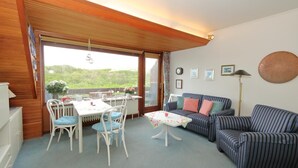 A cozy living room with a dining area, a striped sofa, a table with chairs, and a view of greenery outside. - App. Feusser - at Wenningstedter Strand (Wenningstedt-Braderup (Sylt))