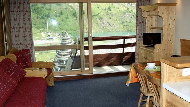 A room with a bed, a dining table with chairs, a TV, and a view of a lake and mountains. - Large studio for 4 - Comfortable with balcony (Tignes)