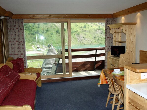 Ein Zimmer mit einem Bett, einem Esstisch mit Stühlen, einem Fernseher und Blick auf einen See und Berge.